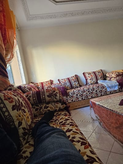 This image depicts an indoor living room within a residence located in Taza, Morocco. A person is present, with their legs visible in the foreground, clad in blue jeans and black socks, reclining on a traditional Moroccan-style sofa (seddari). The sofa is upholstered in patterned fabric featuring floral and geometric motifs in shades of maroon, red, brown, beige, and gold, and includes numerous matching throw pillows, some with maroon fringe. The sofa base exhibits decorative carved wood with gold accents. In the center of the room, a low, rectangular coffee table is situated, covered by a multi-layered patterned tablecloth in orange/red and lighter tones, supported by a metallic base. The room features light-colored walls and a white ceiling adorned with intricate decorative plaster molding. A window on the left side is partially obscured by sheer, multi-colored curtains in orange, yellow, and maroon hues. The floor is covered with light-colored square ceramic tiles. A folded white or light-colored blanket is placed on the far right section of the sofa.