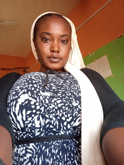 An adult female subject, appearing to be of Nigerian descent, is depicted in a chest-up portrait, looking directly at the camera. She wears a light-colored headscarf, a black long-sleeved garment, and a dark blue or black sleeveless dress with a white, abstract, mottled pattern. A black drawstring is visible at the neckline of her dress, and a black band cinches the waist. Small, gold-colored hoop earrings are visible. The background consists of an interior wall painted in two horizontal sections: the upper section is orange, and the lower section is green, with a visible horizontal dividing line. A piece of paper with unreadable text is affixed to the green wall. The ceiling is a light brown color, and a partial view of a fluorescent light fixture is visible in the upper left background. The image does not provide any visual information to determine the specific location within the city of Muhammadu Dikko Stadium, Nigeria.