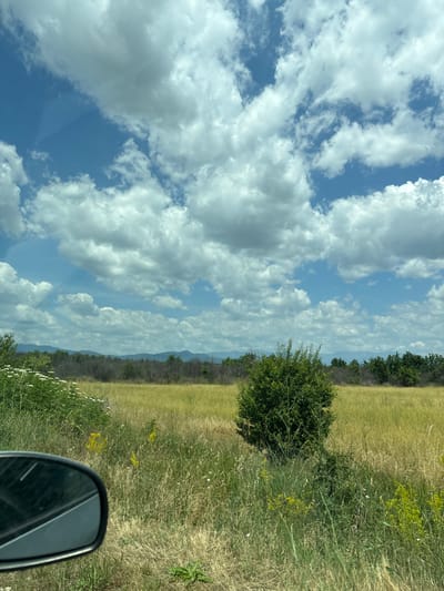 The image depicts a scenic view of a rural landscape, likely in or around Novo Zhelezare, Bulgaria. The foreground features the side mirror of a vehicle, reflecting a portion of the sky. Beyond the mirror, a field of dry, golden grass stretches into the distance, dotted with scattered green vegetation and some yellow wildflowers. A solitary green bush stands prominently in the middle ground. In the background, a line of trees forms a dark border, beyond which a range of hazy blue mountains is visible under a bright blue sky filled with dramatic white cumulus clouds. The overall impression is one of a sunny, possibly summer day in the Bulgarian countryside.