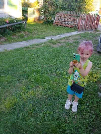 A young child with pink hair is standing on a grassy lawn, holding a smartphone. The child is wearing a lime green tank top and blue shorts, with white sneakers. A small black purse with a gold chain strap is slung across the child's chest. The background shows a wooden fence, some trees, and a building. The scene appears to be outdoors in a residential area.