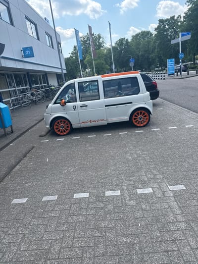 The image depicts a "Mia Amore" vehicle, colored white with orange accents, parked on a street with gray brick pavement, possibly in Veghel, Netherlands, based on the presence of a building with a blue "uw" logo, which is associated with a local financial institution. The vehicle is positioned next to a bike rack filled with bicycles. In the background, there are flags, street signs, and trees under a cloudy sky. Another vehicle is parked behind the Mia Amore. The image is taken from a low angle, showing the building and sky in the background.