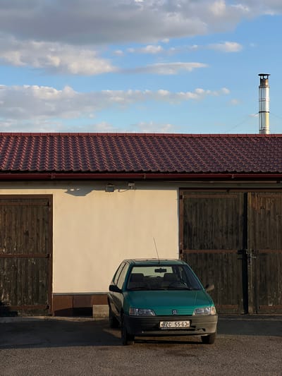 In Újezd u Brna, Czechia, a green Peugeot 106 is parked in front of a building with a red tiled roof. The building has a cream-colored facade and two large wooden doors. A metal chimney extends from the roof on the right side of the building. The sky is partly cloudy, with some cumulus clouds against a blue background. The scene is lit by the warm light of what appears to be late afternoon or early morning.