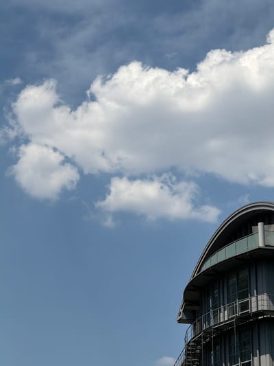 This media file is a photograph taken from a low angle, looking up at a clear blue sky with scattered white clouds. A modern, curved building with reflective glass windows and dark framing is visible in the upper right portion of the frame. The building appears to be an office building or a similar commercial structure. The sky suggests it is daytime, likely mid-morning or afternoon, with good visibility. The clouds are fluffy and white, indicating fair weather. There are no people or animals visible in the frame. The image composition focuses on the interplay between the man-made structure and the natural elements of the sky. The location is suggested to be Frankfurt, Germany, which is consistent with the modern architectural style.