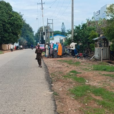 The image captures a street scene in Unguwar Rimi, Nigeria, with an overcast sky. A young boy in a dark traditional outfit is walking towards the camera on the right side of the asphalt road. In the distance, on the left side of the road, several people are gathered near parked vehicles and a small structure. Among them, a man in a white shirt and dark pants is standing, facing away from the camera, appearing to be involved in some activity. Further down the road, other individuals are visible, including a person on a motorcycle. The environment includes trees lining the left side of the road, and a patch of grass and dirt on the right, with a rudimentary outdoor shelter and cooking area. A modern building with signage is visible in the background on the right.
