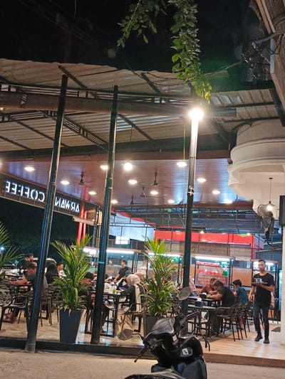This media file depicts an outdoor seating area of a cafe at night, likely in Pantegajah, Indonesia. Several people are seated at tables, engaged in conversation or looking at their phones. A waiter in a black t-shirt is seen walking with a tray of drinks, suggesting food service is ongoing. The cafe features an outdoor covered patio with tables and chairs. Potted plants are strategically placed to enhance the ambiance. Overhead, strings of lights and ceiling fans are visible, indicating a warm climate. A prominent sign with illuminated white letters reads "COFFEE" in reverse, and another sign, also in reverse, shows "NAWIRA". Further back, a menu board displays text, including "NASI GORENG SEAFOOD", "NASI GORENG", and "THE KAH". The scene is illuminated by artificial lighting from the cafe's fixtures and streetlights, creating a warm and inviting atmosphere. A scooter is parked in the foreground, adding to the urban setting. The overall impression is one of a bustling evening at a popular local eatery.