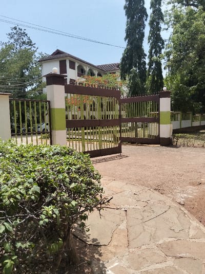 The image depicts the entrance to a property in Waa Ngombeni ward, Kenya, under clear daytime conditions. A partially open, two-leaf metal gate with vertical light green bars and horizontal brown crossbars is central to the midground. The gate is affixed to square pillars, each white with a horizontal light green stripe and topped by a spherical white fixture. Beyond the gate, a white multi-story building with a brown tiled hip roof and brown window shutters is visible. The building features arched openings on its facade.
In the immediate foreground, to the left, a dense, green, leafy bush obscures the base of the gate structure. The ground surface consists of irregular, light-brown flagstone paving transitioning into reddish-brown dirt and gravel. Shadows are cast from the gate and foreground vegetation, indicating bright sunlight.
Various trees populate the scene, including two tall, slender conifer-like trees situated to the right of the building, and broader-leaf trees visible on both sides and behind the building. One tree behind the gate exhibits clusters of small orange-red blossoms. A white vehicle is partially visible through the left side of the gate. Overhead utility wires stretch across the upper portion of the frame against a clear blue sky. No individuals are present in the scene.