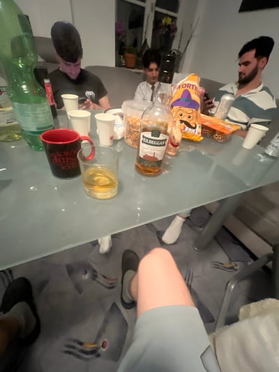 The scene captures an indoor gathering, possibly a casual social event. Four individuals are partially visible around a reflective, likely glass-topped table. One man, sporting a beard and a striped shirt, is observed using a mobile phone. A second man is also present, though his actions are unclear. Two additional individuals are visible primarily from the waist down, wearing shorts and socks.

The table is laden with items indicating food and drink consumption. Objects include a bottle of Kilbeggan whiskey, other alcoholic beverages, cups (some filled), and snacks such as tortilla chips and pretzels.

The environment appears to be a residential living space, with a sofa partially visible in the background. The scene lacks specific details to identify its location within Benningen, Germany.