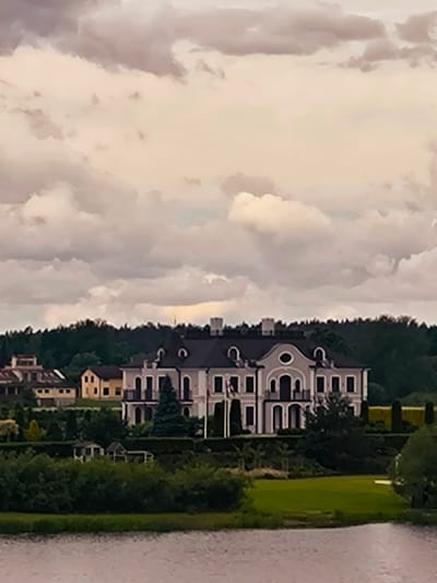 This is a vertical, slightly blurred, medium-range photograph of a grand, multi-story house situated on a hill overlooking a body of water. The house is light gray with a dark roof and features numerous arched windows, balconies, and decorative elements. It is surrounded by lush green trees and manicured lawns. In the background, a dense forest lines the horizon. Above the landscape, a dramatic sky filled with heavy, overcast clouds dominates the upper portion of the image. The body of water in the foreground appears calm with subtle ripples. The location is identified as the city of Salaspils, Latvia, and the house is part of a residential area with other, smaller houses visible in the distance to the left. There are no people visible in the scene.