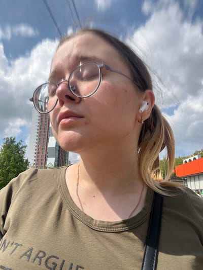 A close-up selfie of a woman wearing glasses, an AirPod in her ear, and an olive-green t-shirt with partial text reading "N'T ARGU". The background includes a cloudy sky, a building, and trees. The building with many windows is near Serpukhov, Russia.