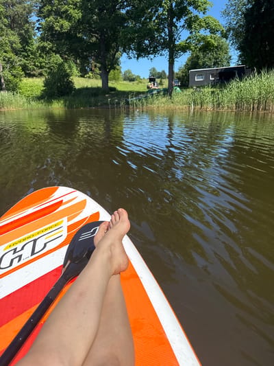 A first-person perspective image taken from a paddleboard on a body of water. The paddleboard is orange and white with the brand name "HYDRO-FORCE" visible. A person's bare legs and feet are resting on the paddleboard, with a paddle lying next to them. The water is dark and reflects the surrounding trees and sky. In the background, a wooded shoreline with tall reeds is visible. A small, dark-colored building with windows and an open entrance is situated among the reeds. Further back, a grassy hill rises, dotted with trees. The sky is clear and blue. The location is identified as Lendži, Latvia.