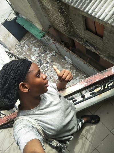 A young dark-skinned female is depicted on a tiled balcony in Cotonou, Benin. She has black braided hair with a black headband and wears a light grey short-sleeved t-shirt with a dark logo on the left chest, a black stud earring in her left ear, patterned shorts or pants, and a black slipper on her left foot. A light-colored woven strap bag is carried over her right shoulder. She is positioned behind a rusted metal balcony railing with ornate scrollwork, looking towards her right (viewer's left) with her mouth slightly open, while giving a 'thumbs up' gesture with her raised right hand. The balcony floor consists of light grey square tiles, partially shaded. Below, a ground-level courtyard is visible, paved with fragmented tiles and concrete. Adjacent structures include a light-colored wall to the left, with a dark green corrugated metal awning and a dark opening below. To the right, a multi-story building made of dark grey blocks features reddish-brown boarded windows and a weathered grey corrugated roof. Barbed wire is affixed to a lower wall or barrier separating the ground area from this building, and white fabric hangs below the windows.