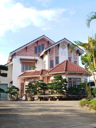 This image captures a large, multi-story residential building in Đa Thọ, Vietnam, presented under a bright blue sky with scattered white, wispy clouds. The house features a complex architectural design with multiple steeply pitched gabled roofs, all covered in reddish-brown terracotta tiles. The exterior walls are primarily a light, possibly cream-colored, stucco or painted finish, contrasted with reddish-pink exposed brickwork on the upper sections of the gables. Numerous wooden-framed windows, mostly tall and narrow, are distributed across the facade; a large bay window is prominent on the lower right. Decorative elements include dark-colored support brackets beneath roof eaves and window awnings, and a circular design with horizontal banding on the central gable. A lightning rod is visible on the peak of the leftmost roof.
The foreground consists of a wide, light-grey paved area composed of rectangular pavers, displaying distinct shadows indicative of direct sunlight. This area leads to a meticulously landscaped section directly in front of the house, featuring numerous large, well-shaped bonsai trees arranged in decorative ceramic pots and on substantial stone tables. To the far left, a light green roll-up garage door is partially visible. On the far right, a section of a palm tree's fronds extends into the frame, and a cultivated garden bed with a variety of colorful flowering plants (pinks, purples, and oranges) is present. No individuals are visible within the scene.