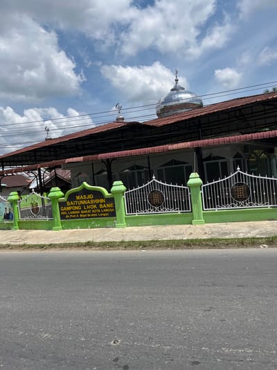 The image captures the facade of the Baitunnashihin Gampong Lhok Banie Mosque, located in the Langsa Barat subdistrict of Langsa City, Indonesia. The mosque features a prominent silver dome topped with a crescent moon and star symbol, indicative of Islamic architecture. The building itself has a reddish-brown tiled roof supported by black metal beams, with a white and green fence enclosing the front of the property. A green signboard with the mosque's name and address is prominently displayed. The street in front of the mosque is paved with asphalt, and there is a strip of grass and concrete bordering the road. The sky is partly cloudy with a clear blue sky visible between white cumulus clouds. No people are visible in the frame.