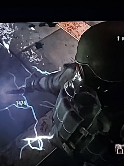 The image is from a video game and depicts a first-person perspective. The player character, a soldier wearing a helmet and body armor, is in an urban environment. The player character is holding a weapon, presumably a rifle, and is looking down at a patch of ground. The ground is illuminated by a bright blue light, possibly from an electrical source, and a number "1474" is visible in the foreground. The environment is dark and gritty, suggesting a war-torn or post-apocalyptic setting.  The coordinates provided, 49.201, -122.678, correspond to a location in the city of Pitt Meadows, Canada, however, the image does not reflect any details of this location.