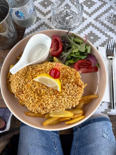In Ravda, Bulgaria, on a table covered with a gray and white geometric patterned tablecloth, a plate of food is centrally located. The plate contains a large, breaded cutlet, several french fries, a small salad, and a garnish of a lemon wedge and a cherry tomato. To the left of the plate, a white ceramic gravy boat holds a white sauce, and a partially filled 1-liter bottle of water is visible. To the right of the plate, a fork rests on a white napkin. The scene is viewed from a high angle, with the lower portion of the image showing a person's lap wearing blue jeans with ripped details. A crystal glass is also present on the table.