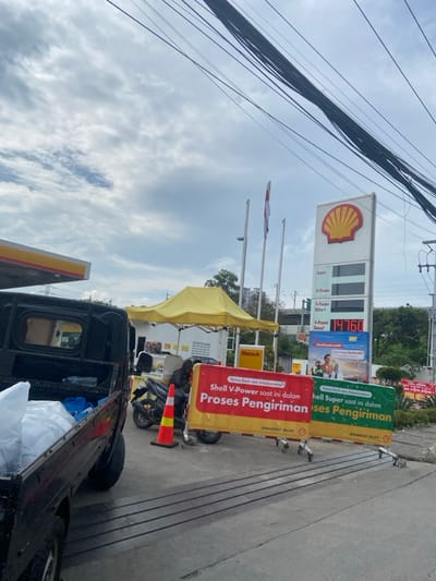 The image shows a gas station in Bojongsoang, Indonesia, located at coordinates -6.967 latitude and 107.638 longitude. The scene is a gas station with a black truck parked at the side, with a large sign displaying "Shell V-Power saat ini dalam Proses Pengiriman" and "Shell Super saat ini dalam Proses Pengiriman" in front of it. The sign is red with yellow borders and a white background, and is placed on the ground. The truck is parked at the side of the gas station, with its bed filled with white and blue blankets. There is a person sitting on the back of the truck, seemingly waiting. Behind the sign, there is a small yellow canopy tent. The scene is illuminated by sunlight, and the sky is clear with some clouds. There are a few power lines overhead. There is a large Shell sign, with a digital display showing the price of fuel, on the right side of the image. To the left of the Shell sign, there is a small white flagpole. The image focuses on the signs and the truck, with the gas station in the background. The scene appears to be a typical gas station in Indonesia, with a clear focus on the Shell brand and the process of sending fuel. The image seems to be taken during the day, with no indication of specific time of day.