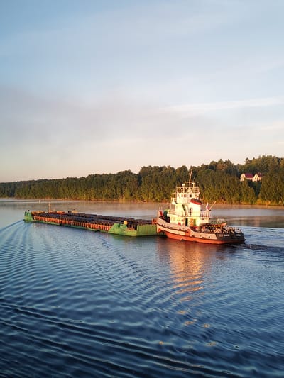A tugboat and an attached barge navigate a wide body of water in Kabanovo, Russia, during what appears to be either sunrise or sunset. The tugboat, primarily white with a red hull, displays the markings "ЩВЛ30" and "ТСН-41" on its starboard side and flies a small flag. It is positioned to the right of the barge, pushing it forward, creating a distinct rippled wake in the calm, dark blue water. The long, green-hulled barge is heavily laden with numerous dark, cylindrical objects, likely industrial tires or similar large rubber items, stacked in organized rows across its deck.
The sun's low angle illuminates the scene with warm, golden light, casting reflections on the water surface and the vessels. The far bank of the waterway is densely covered with lush green trees, creating a continuous forested shoreline. On a rise within this forest, towards the right side of the image, two small, white houses with prominent red roofs are visible. In the water, closer to the far bank and to the left of the barge, a white conical navigational buoy is present. The sky above is clear, transitioning from a light blue at the zenith to a pale orange-pink hue near the horizon. No individuals are visible on either vessel or along the shoreline.