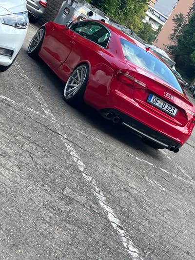 A red Audi A5 coupe is parked on a paved lot with white painted parking lines in Dietzenbach, Germany. The car is facing away from the camera and is angled to the right. It has a license plate that reads "OF D 323". The car is clean and appears to be in good condition. The background includes a building and trees. The weather appears to be sunny. The lot is likely a public parking area.