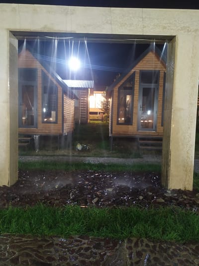 In the city of Sosnovka, Kyrgyzstan, at night, a view is presented through a concrete archway which has water spraying downwards from the top of the arch. In the background, two small, wooden, cabin-like structures with pitched roofs and dark-trimmed windows and doors are visible. The structures are illuminated by a bright light, possibly a streetlamp, positioned above and between them, casting a strong glare. To the left of the cabins, a light emanates from a building. The ground in front of the cabins consists of green grass and an area covered with dark, wet stones and debris, also impacted by the falling water. A small garden gnome is visible on the grass to the right of the left cabin. The overall scene suggests an outdoor recreational area or a glamping site in inclement weather.