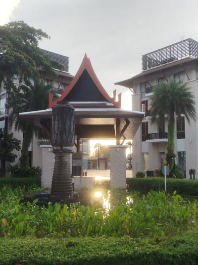 The image depicts the exterior of a building complex, possibly a hotel or resort, in Ko Kaeo, Thailand. In the foreground, a body of water is filled with lush green aquatic plants and smaller, light-colored flowers. The water reflects the bright sunlight, creating a strong glare. A well-maintained hedge lines the bottom of the frame. Dominating the mid-ground is a decorative pillar or sculpture, dark in color with a textured, patterned surface. Behind the pillar, a traditional-style roof structure with a pointed, dark gable and reddish-orange trim is visible. This structure appears to be an entrance or pavilion.

The background features a modern multi-story building with a light-colored facade, balconies, and numerous windows. Palm trees, including one with dense foliage growing up its trunk, are present. A sign with green text on a white background is affixed to the building on the right side. The sky is overcast with some brightness indicating sunlight filtering through. The overall impression is of a tropical setting with a blend of traditional and modern architectural elements. There are no people or animals visible in the image.