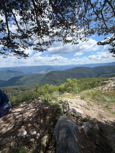 A scenic vista unfolds from a rocky outcrop under the dappled shade of pine trees. The foreground shows the tire and mudguard of what appears to be a dirt bike, suggesting an off-road adventure. To the left, a blue and white patterned piece of fabric, possibly part of a rider's gear, is visible. The midground presents a lush, undulating landscape of green, forested hills and valleys. In the far distance, softer, hazy blue mountains recede into the horizon. The sky above is a bright blue, punctuated by fluffy white clouds, indicating a clear, sunny day. The exact location within Kosovo, Bulgaria cannot be determined from the visual information provided.