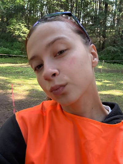 The media file shows a close-up, Dutch angle shot of a young person outdoors. The subject has light skin, light brown hair, and is wearing an orange vest over a black garment. They are also wearing blue-tinted eyeglasses on their head. The background is blurred but appears to be a natural, wooded environment with green grass and trees, suggesting a park or forest setting. Sunlight is filtering through the trees, creating dappled lighting. The time of day appears to be daytime, likely late morning or afternoon, given the brightness and the shadows. The person's expression is neutral or slightly contemplative. There are no visible text elements in the image. The location context provided is Сергино, Russia. The scene suggests an outdoor activity or event, possibly a sporting event or a casual outing in nature, given the vest and the outdoor setting.