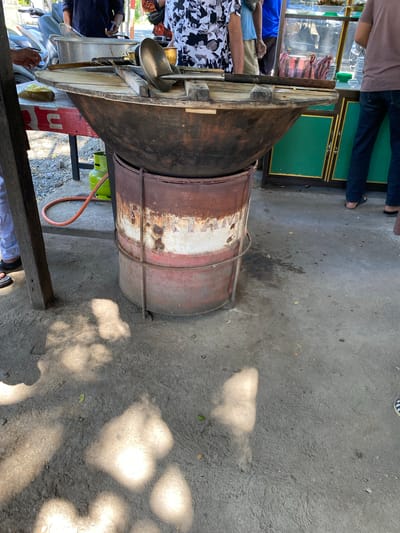 In Meunasahmanyang, Indonesia, a large, round, blackened cooking vessel, likely a wok or cauldron, sits atop a rusted metal barrel that has been repurposed as a stand. The barrel displays faded red and white bands and is held together by wire. A green propane tank with an orange hose is visible behind the setup. The scene is outdoors, with a concrete ground surface casting dappled shadows from overhead foliage. A wooden table with a red and white striped cloth is partially visible to the left, holding a yellow plastic-wrapped item and some cooking utensils, including a large metal scoop. Several individuals are present, with parts of their legs and torsos visible, suggesting they are involved in food preparation or service. To the right, a green and yellow display cabinet, possibly for food, is also in view. The overall impression is of an outdoor food stall or a cooking area in a public space.