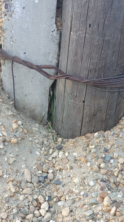 This is a close-up, outdoor, eye-level photograph focusing on the base of a weathered wooden post and a section of concrete structure, surrounded by sandy soil and small pebbles. The wooden post, which appears to be a utility pole, has a rough, textured surface with prominent vertical grain and some dark markings, possibly insect holes or knots. A bundle of rusty, twisted metal wires is wrapped around the post and extends towards the concrete. The concrete structure is a rough, unpainted surface with some visible aggregate and a crack running vertically. Between the wooden post and the concrete, a narrow gap allows a glimpse of green grass growing. The ground is covered with a mixture of sand and various sized, rounded pebbles in shades of gray, brown, and white. The lighting suggests it is daytime with no strong shadows, indicating possibly overcast conditions or indirect sunlight. The scene is devoid of any human presence or activity.
