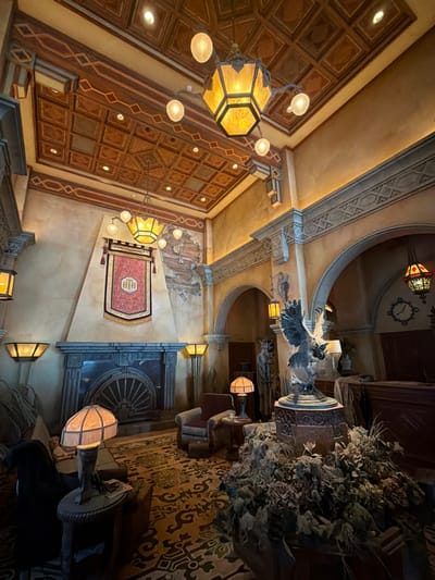This is a wide-angle, upward-facing shot of an elaborately decorated interior, possibly a lobby or grand room. The decor suggests an exotic or historical theme, with ornate wooden paneling on the ceiling and walls, intricate geometric patterns, and what appears to be a tapestry or rug hanging on one wall. Several light fixtures are visible, including a large, multi-bulbed lantern hanging from the ceiling and smaller wall-mounted lamps that cast a warm glow.
In the background, a large, detailed clock face is mounted on the wall. A prominent statue or sculpture of a bird of prey is also visible, perched above an arched doorway. The overall atmosphere is dimly lit and somewhat mysterious, with shadows playing a significant role in the composition. The detail of the wood and textile work suggests a high-quality, possibly luxurious, setting. There are no people visible in the frame, and the scene appears still. The lighting suggests it is either evening or the interior is designed to evoke such an atmosphere. The context of Chessy, France, might imply a themed attraction or hotel. There is no discernible weather information.