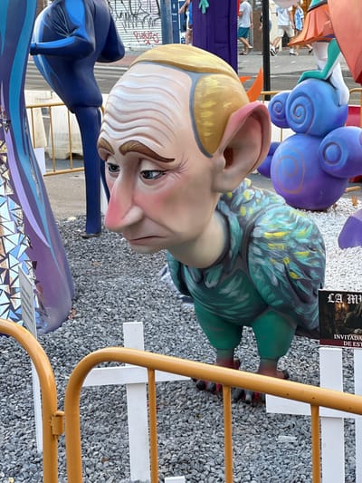 The image depicts a street scene featuring elaborate, oversized figurative sculptures, typical of a Fallas festival, likely in Alicante, Spain. The primary subject is a large, stylized head of a person with exaggerated features, including large ears, prominent nose, and wrinkled skin rendered in pale tones with rosy cheeks. The figure's blond hair is painted, and its eyes are a piercing blue.
Behind and to the left of the main head, other colorful sculptures are visible. These include a large, coiled, purple shape resembling a cloud or a character, and parts of other figures in orange and blue. The sculpture in the foreground, on the right, is a torso covered in a green and yellow painted patterned fabric, with red accents on its feet.
The sculptures are set against a backdrop of gravel and a white picket fence, with a bright yellow metal railing in the foreground. In the background, parts of other figures and structures can be seen, as well as some people observing the displays. The lighting suggests it is daytime, possibly sunny, casting shadows.
A small sign is visible attached to the green sculpture, featuring some text in what appears to be a gothic font. The visible text reads "LA MAN" followed by "INVITADA DE EST". The overall impression is one of a festive, public display of art and cultural tradition.