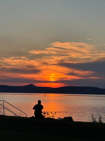 At sunset, a man stands silhouetted at the shoreline of Lake Balaton, Hungary. His posture suggests he is fishing, near rods supported by a rod stand. The sky contains orange-hued clouds, partly obscuring the sun. The lake water reflects the orange light. A dark treeline forms the horizon behind the lake. A railing runs down a grassy bank in the foreground to the lake.