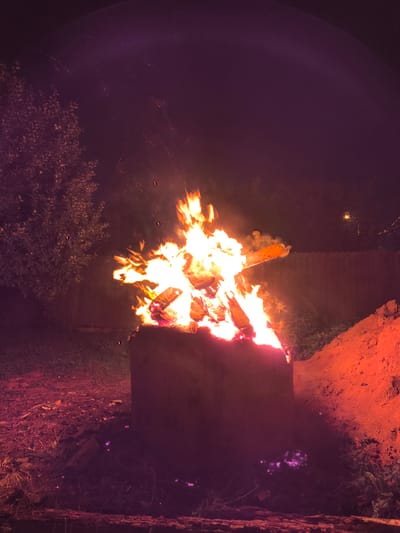 In Bylovo, Russia, a bonfire burns brightly in a concrete fire pit at night. The flames reach upwards, casting a warm orange and yellow glow on the surrounding logs and sparks fly into the dark sky. To the left of the fire pit, a leafy tree is visible, its branches silhouetted against the night. To the right, a pile of reddish-orange dirt rises, illuminated by the firelight. The ground around the fire pit is covered in dry leaves and grass. The scene is framed by a wooden fence in the background. The overall atmosphere is one of warmth and rustic simplicity.