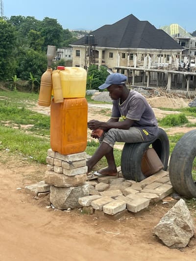 In Galadimawa Village, Nigeria, a person wearing a blue cap and a grey shirt with yellow trim is working. They are crouched down, reaching towards a stack of plastic containers, one yellow and one orange, which are resting on a makeshift base of bricks and tires. A large, weathered tire is positioned behind the containers, and another large tire is visible in the upper right corner of the image.
The background shows a construction site with a multi-story building under development. Several individuals can be seen on the building's upper floors, engaged in construction work. The surrounding area is a mix of dry earth and sparse green vegetation, typical of a developing area. Sunlight suggests it is daytime, and the weather appears to be clear. The person's focused posture and the construction activity indicate a scene of labor and ongoing development. The text ".com" is visible on the yellow trim of the person's shirt.