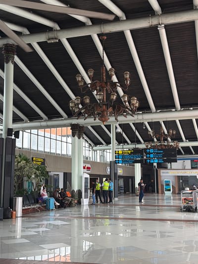 The image depicts the interior of an airport terminal, likely located in Tangerang, Indonesia, given the Indonesian signage. The scene is spacious, with a high, dark wooden ceiling supported by white beams and columns. A large, ornate, multi-tiered chandelier hangs from the ceiling. The floor is highly reflective, tiled with a pattern of light and dark squares, mirroring the ceiling structure and other elements in the terminal.
In the foreground, a group of people are seated on a bench, some with luggage. To the left, a silver trash can is visible, and further left, part of a plant is seen. In the mid-ground, several individuals are standing near signage. Two people in high-visibility vests appear to be airport staff or security, conversing with another individual. To the right, a digital information screen is present, labeled "inJourney AIRPORTS CUSTOMER LOUNGE". A luggage cart is parked nearby.
Multiple directional signs are suspended from the ceiling, indicating various airport services and locations such as "Kedatangan 1B Arrivals 1B", "ATM ATM Center", "Bus Umum Public Bus", and "Toilet Toilets". The overall environment suggests a functional and busy airport concourse.