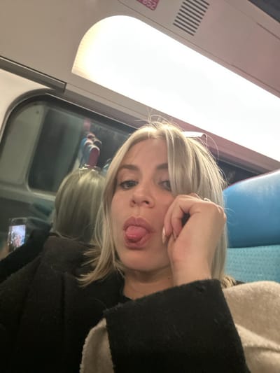 A woman with blond hair poses in what seems to be a train. She has one hand near her forehead, her lips are puckered, and she is sticking out her tongue. She's wearing a dark jacket and a ring on her left ring finger. A window with a reflection is behind her and a bright light is above. The image is slightly angled.