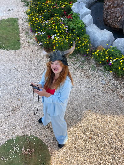 In Thôn 1, Vietnam, a person with long, reddish-brown hair wears a Viking helmet with horns, a red top, and a light blue pantsuit. They are standing on a ground covered with small, light-colored pebbles. The person is holding a black electronic device in their hands and has their tongue sticking out, appearing to be making a humorous expression. To the upper left, a patch of green grass is visible, also scattered with pebbles. In the background, a dense flower bed with numerous yellow, white, and red blossoms is situated next to large, gray, sculpted rocks that form a decorative border.