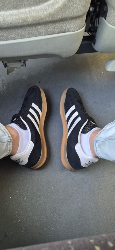 A pair of black Adidas sneakers with white stripes and gum soles are shown from a top-down perspective, resting on a speckled gray floor. The sneakers are worn with white socks and light-colored jeans. The background consists of the textured upholstery and plastic trim of a seat, possibly in a vehicle. The scene appears to be indoors, given the lighting and indoor materials visible. The location in the city of Gaupen, Norway is not discernible from the image.