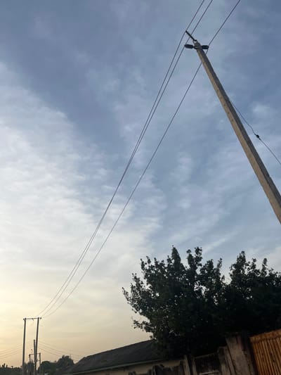 The image captures an outdoor scene in Barnawa, Nigeria, featuring prominent utility infrastructure against a partially cloudy sky. A weathered concrete utility pole, equipped with insulators and multiple dark power lines, dominates the upper right quadrant, extending diagonally towards the lower right. Three main power lines stretch horizontally across the middle of the frame, converging towards the top of the pole. In the lower middle and right, the dark green canopy of a large tree is visible. Below the tree, a dark-colored roofline of a building is partially visible, fronted by a low concrete wall. A rusty, metallic gate is present on the far right, partially obscured by the tree foliage and the wall. In the distant lower left, a second, smaller utility pole with additional power lines is visible against a lighter, hazy sky. The sky transitions from a light blue-grey with scattered white cloud formations at the top to a warmer, lighter hue towards the bottom left, suggesting either early morning or late afternoon conditions. No individuals or specific activities are observable.