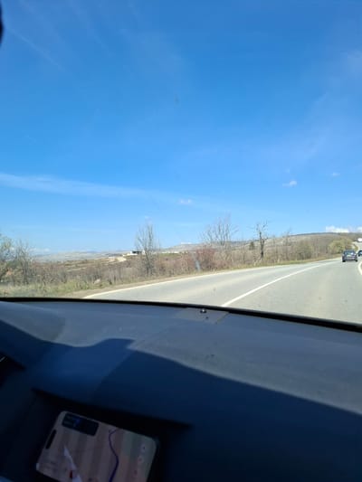 The image depicts a view from inside a vehicle traveling on a road near Gradets, Bulgaria. The sky is clear and blue. A two-lane asphalt road is visible, with a white line separating the lanes. Vegetation and sparse trees line the roadside. Another vehicle is ahead on the road. A smartphone displaying a map is visible on the dashboard. The location can be determined through the geographical reference of Gradets.
