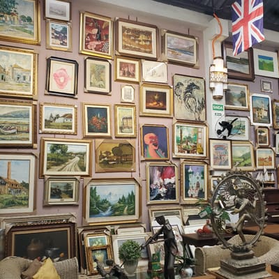 A crowded wall covered with framed pictures in a cluttered commercial space. The walls are a light pink hue, and the paintings vary in size and style, ranging from landscapes to still life to abstract pieces. A Union Jack flag is partially visible in the upper-left corner, along with a light fixture and a small, black fan. A green sign with the word "SAIDA" and an arrow is also visible. In the foreground, part of a decorative metallic object and other decor items, including wicker baskets and possibly statues, obscure some of the pictures. The scene is in Curitiba, Brazil.