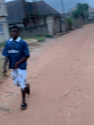 A blurry, sideways shot shows a young man standing on a dirt road. He wears a dark blue shirt and white shorts, along with black sandals on his feet. Buildings with concrete walls stand in the background, partially obscured by vegetation. The sky is overcast, lending a muted tone to the scene. The overall image quality is poor.