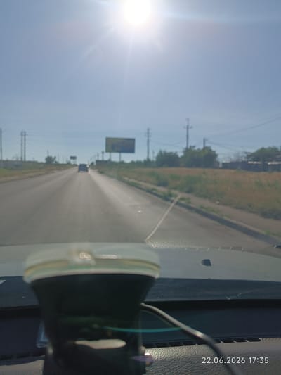 The image captures a view from inside a vehicle driving down a straight road on a bright, sunny day. The sun is high in the sky, causing lens flare and overexposing the upper portion of the image. The road appears to be in good condition with clear lane markings. Vegetation lines the sides of the road, with some trees and bushes visible in the distance. There are power lines and utility poles along the roadside, typical of a rural or semi-rural setting.

A billboard is visible in the distance on the right side of the road, though its advertisement is unclear due to the distance and blurriness of the image. A dark-colored car is in front of the vehicle, further down the road. The foreground shows a portion of the vehicle's dashboard and a device mounted on the windshield, possibly a dashcam or GPS. The device has a white and green casing.

The lower right corner of the image contains a timestamp: "22.06.2026 17:35", indicating the date and time the photo was taken. The weather appears to be clear and warm, suggested by the bright sun and clear sky. The overall scene depicts a typical daytime drive on a highway or main road in Ukraine, specifically noted as Lymanka. No people are clearly visible in the image. The mood is one of travel and everyday activity.