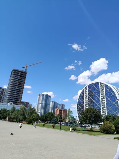 This is an eye-level, outdoor, medium shot of a city park in Grozny, Russia, on a clear, sunny day with scattered clouds. In the foreground, a paved area with a grey cobblestone pattern is visible. Two individuals are in the mid-ground: a person in a white shirt and dark pants is crouching on the left, seemingly taking a photograph, while a person in a light blue dress walks away from them on the right. Further back, a well-maintained lawn area is dotted with trees and bushes, and a lamppost stands on the right edge of the park. Behind the park, the cityscape features several modern buildings. On the left, a tall, unfinished skyscraper with a yellow construction crane extending from its top is under construction. To its right, several other apartment buildings are visible. Dominating the right side of the frame is a distinctive circular building with a blue, diamond-patterned glass facade. Several cars are parked along the street bordering the park.