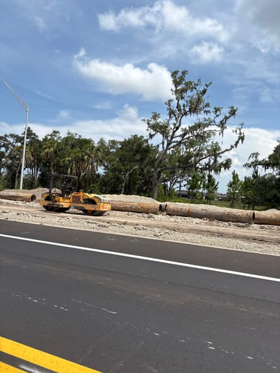 The image shows a roadside construction scene in Parrish, United States. A yellow Volvo roller, a piece of heavy machinery used for compacting surfaces, is parked on the side of an asphalt road that is partially constructed. The road has a white line marking its edge, and a double yellow line is visible on the right side of the image.
The background features lush green trees, including palm trees and mature oak trees draped with Spanish moss, suggesting a warm, humid climate. The sky is a bright blue with scattered white clouds, indicating it is daytime, likely late morning or early afternoon. In the distance, hints of houses can be seen through the trees, suggesting a semi-rural or suburban setting.
The construction activity is evident from the roller, the exposed soil and gravel beside the road, and what appear to be sections of large pipe laid along the embankment. The scene appears peaceful and still, with no people visible. The overall impression is one of ongoing infrastructure development in a natural, sun-drenched environment.