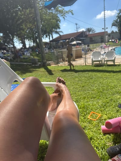 In Beit Yitzhak-Sha'ar Hefer, Israel, a person is seen from the thighs down, reclining in a white lounge chair on a grassy lawn. Their tanned legs are visible, with their feet resting on the grass. In the background, several other white lounge chairs are set up, some occupied by people. A blue swimming pool is visible with a fence surrounding it. Further in the background, buildings and trees are present under a clear blue sky. The scene appears to be a public pool area on a sunny day.