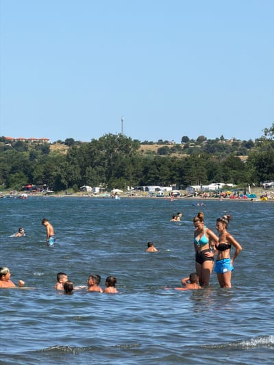 In Atiya, Bulgaria, a sunny day at the beach is depicted with clear blue skies and calm, dark blue water. The foreground shows several people wading and swimming. In the middle ground, a group of children and adults are enjoying the water, with some children playing together. Further back, along the shoreline, a sandy beach is lined with trees, tents, and recreational vehicles, suggesting a campground or a bustling tourist area. A communication tower is visible in the distance, atop a hill covered with vegetation and dotted with houses. The overall scene conveys a lively and relaxed atmosphere of a popular summer destination.