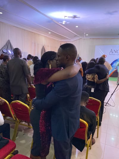 The image depicts a group of people in what appears to be an indoor event, possibly a religious gathering, in Miango, Nigeria. The focus is on a couple embracing; the man is wearing a dark blue suit, and the woman is in a dark dress with red sequin details and a maroon fur-like scarf. They are standing close, with the man's arm around the woman. Other individuals are seen in the background, dressed in various formal attires, some seated on red and gold chairs. A banner with the word "ANOINTING" and a scripture reference "MATTHEW 7:24" is visible in the background. The environment suggests a hall or large room with white walls and ceiling, equipped with ceiling fans. There's also lighting equipment visible, indicating the event is planned and potentially staged.