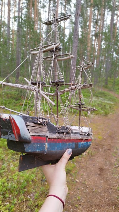 A hand with grey nail polish and a burgundy bracelet holds a weathered model ship. The ship is detailed with masts, rigging, and a hull painted blue and red with some damage. The model is positioned in a natural outdoor setting. The background consists of a forest with tall, slender trees and green undergrowth. A dirt path is visible winding through the trees. The lighting suggests it is daytime, with a slightly overcast or diffused light. The overall scene conveys a sense of rustic charm and perhaps a touch of nostalgia. The location is identified as Orekhovo, Russia.