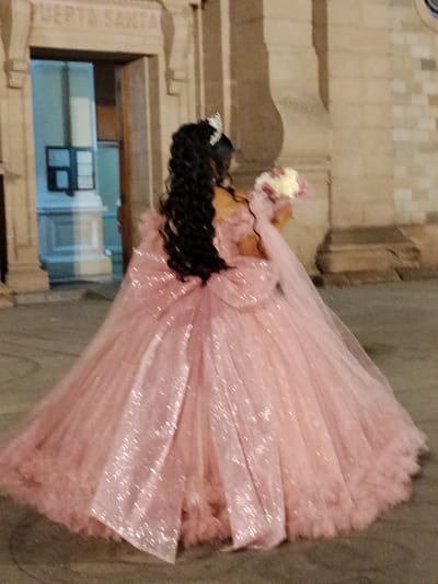 A young woman in a large pink quinceañera-style gown stands outside a building in Tacna, Peru. The gown has a large bow at the back and is heavily layered with tulle. She is wearing a tiara and holding a bouquet of flowers. The building behind her is stone with a sign that reads "PUERTA SANTA." The lighting suggests it is evening or late afternoon.