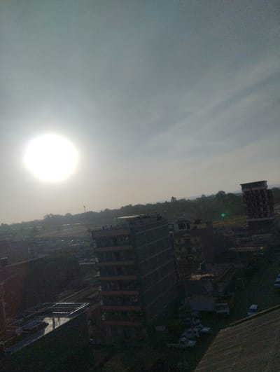 An elevated view captures a cityscape under a bright, hazy sun. Tall buildings dominate the scene, one with scaffolding-like exteriors. Shadows cast across the landscape hint at the early morning or late afternoon light. Vehicles are visible near the buildings. Distant trees and hills form a soft horizon, contrasting with the urban structures.